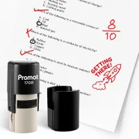 Black self inking stamp and removable cap displayed beside a graded science worksheet marked with red check marks, an “8/10” score, and a rocket stamp that says “GETTING THERE!” The worksheet includes multiple choice science questions, and this ink stamp