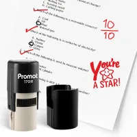 Black self inking stamp and removable cap displayed beside a graded science worksheet marked with red check marks, a “10/10” score, and a smiling star stamp that says “You’re A STAR!” The worksheet includes multiple choice science questions, and this ink